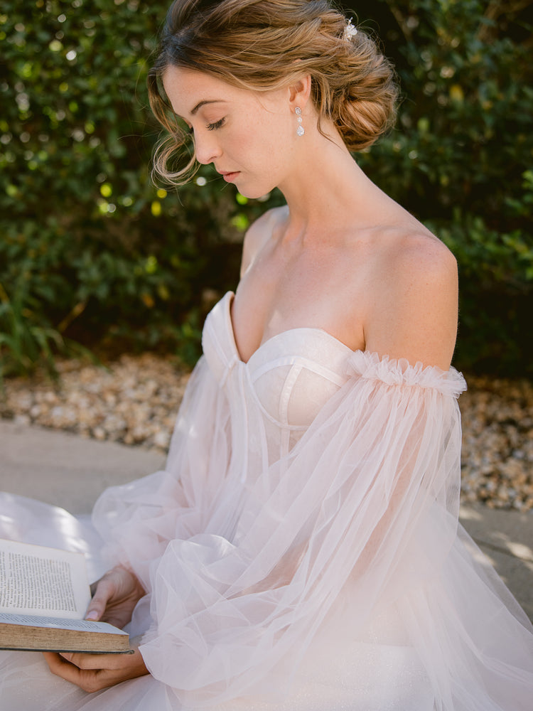 bride reading a book in a blush tulle wedding gown and wearing silver crystal long drop earrings with romantic updo and silver headpiece